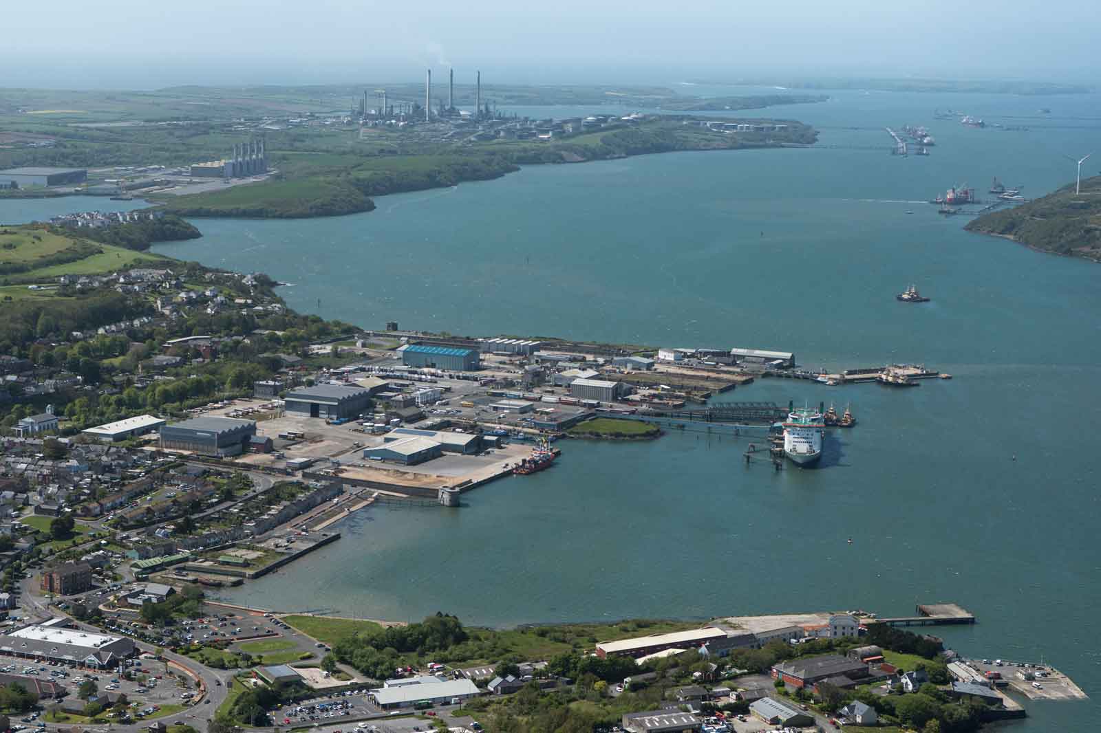 Aerial view of a busy port with industrial buildings, boats in the water, and green hills in the background.