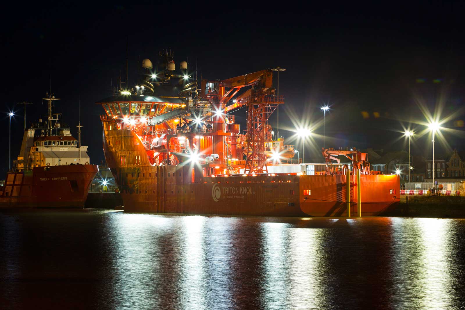 A brightly lit ship named Triton Knoll docked at night, reflecting on the water alongside another vessel, Shelf Express.