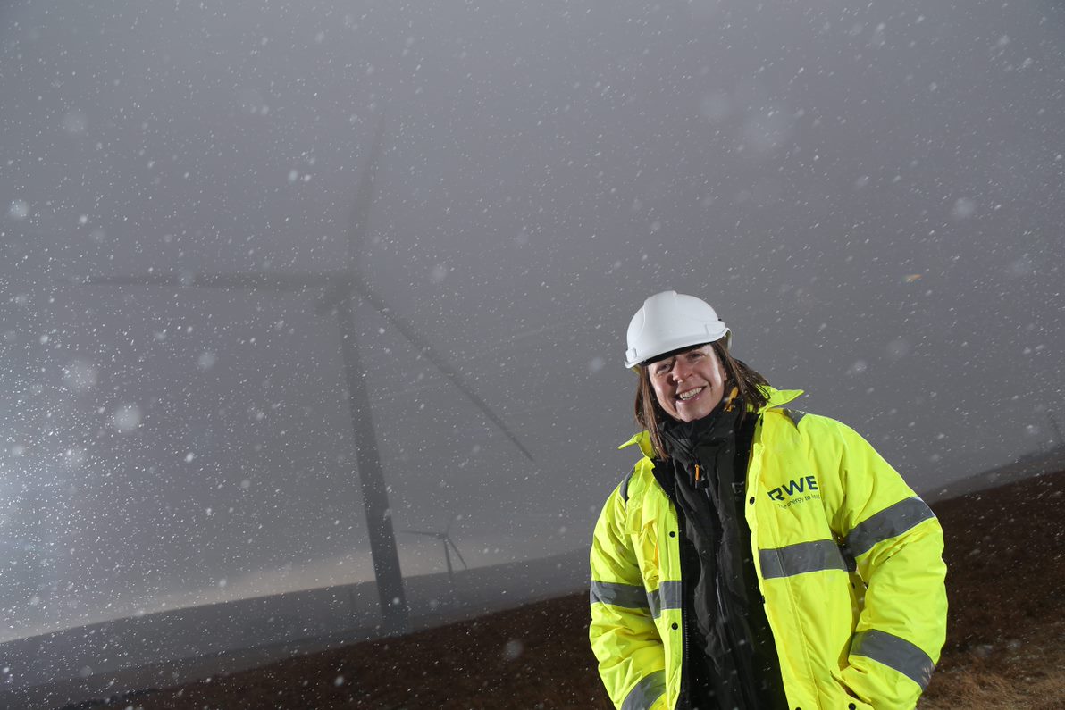 A worker in a bright yellow jacket and white hard hat stands in snowy conditions near a wind turbine.