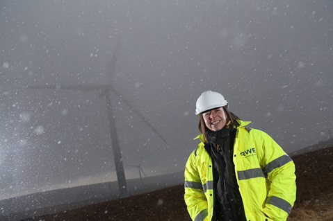 A worker in a bright yellow jacket and white hard hat stands in snowy conditions near a wind turbine.