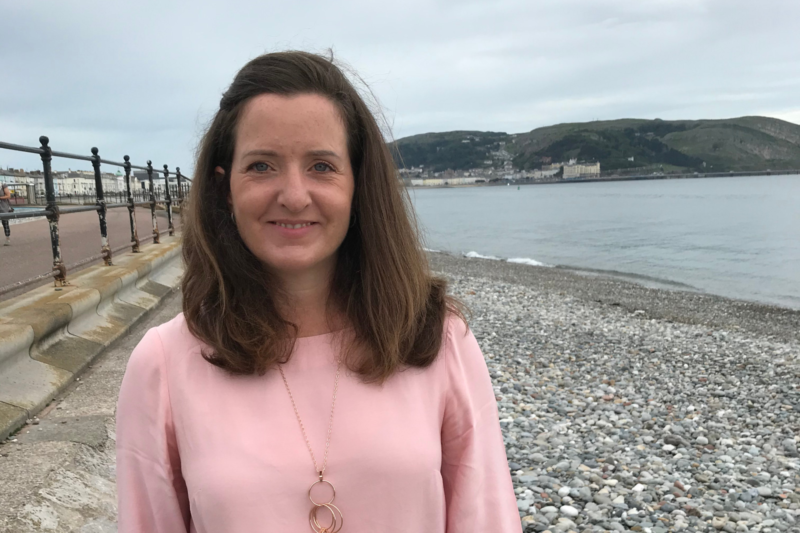 A woman stands on a pebble beach wearing a pink top, with a coastal landscape in the background.