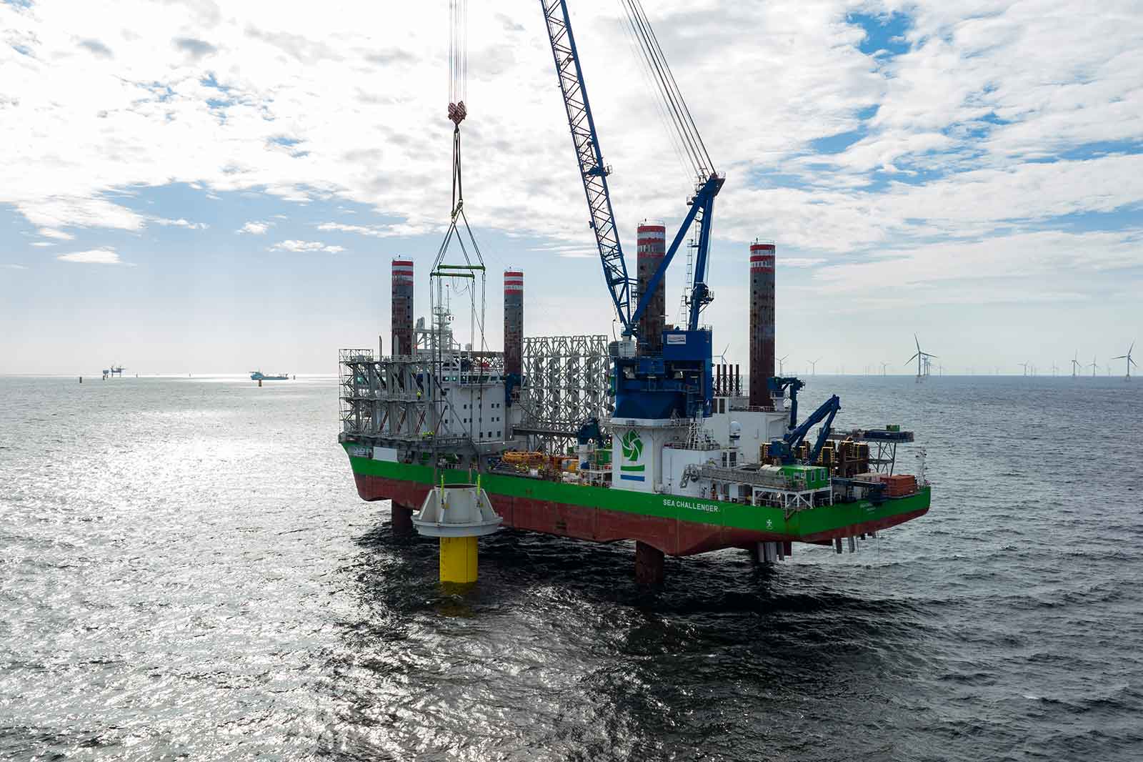 A work vessel with cranes is installing a structure in the sea. Wind turbines can be seen in the background.