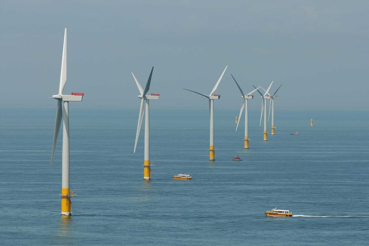 A row of offshore wind turbines stands in calm waters, with boats navigating nearby.