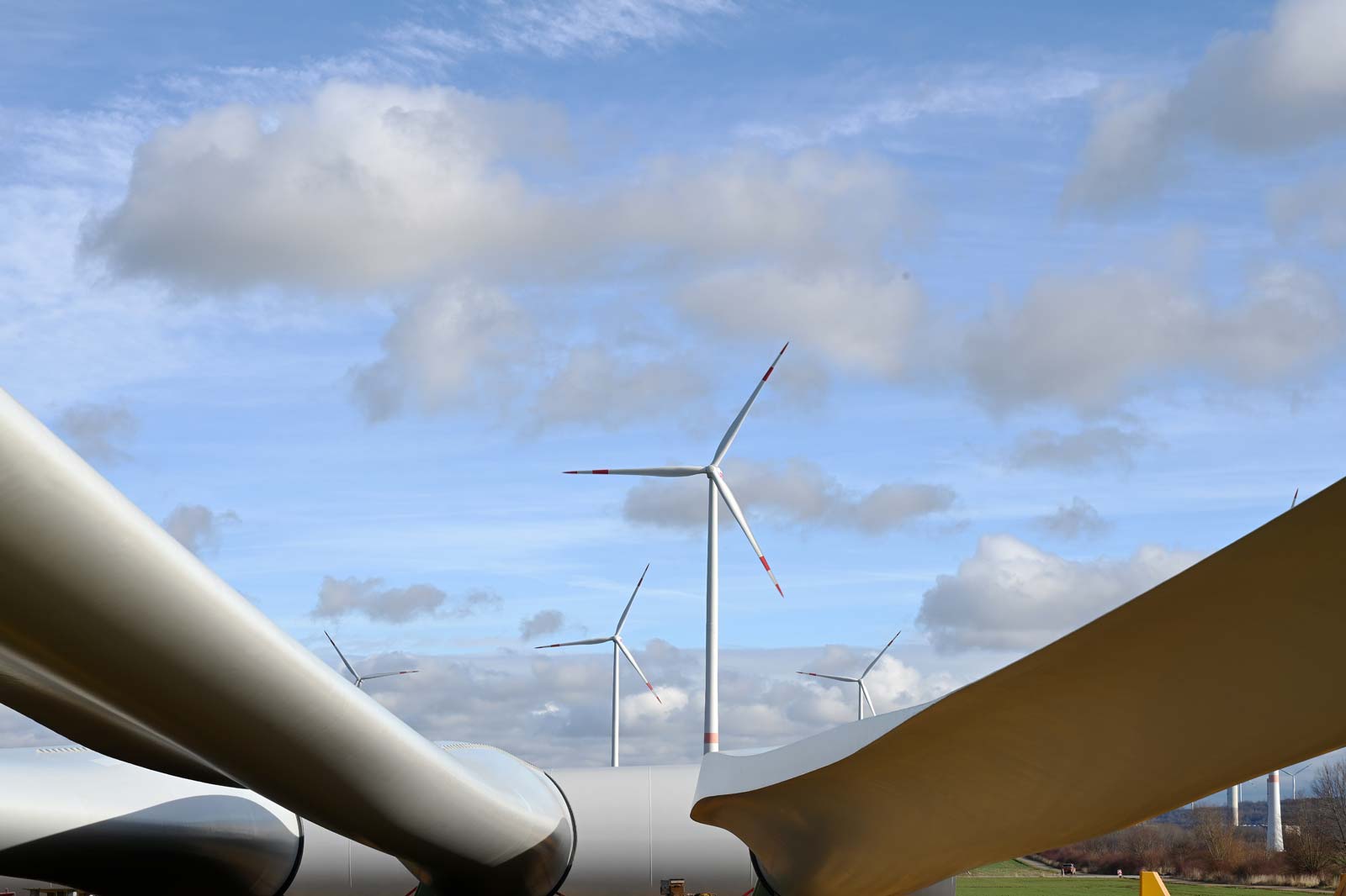 Wind turbine blades against a cloudy blue sky with multiple wind turbines in the background.