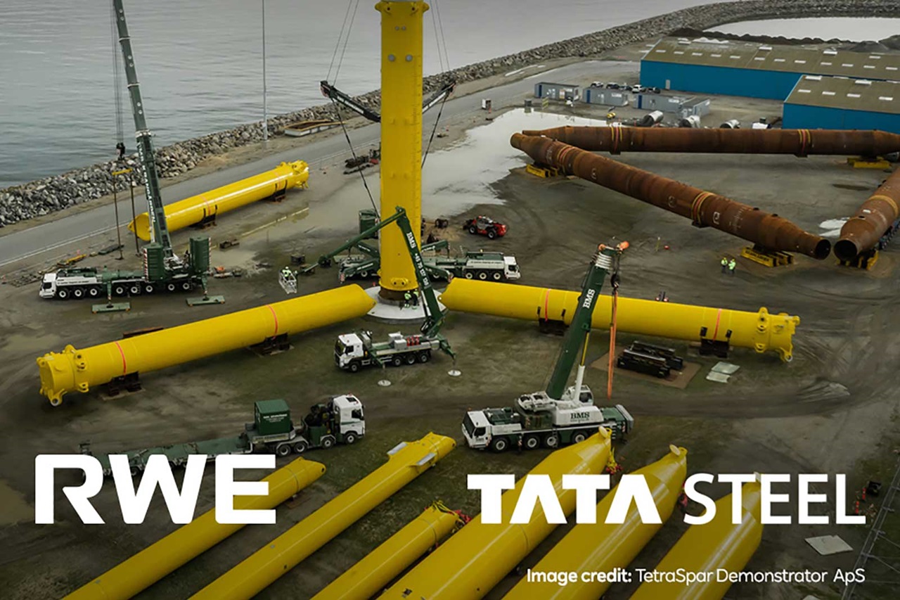 An aerial view of a construction site with yellow cylindrical structures, cranes, and trucks, indicating offshore wind turbine preparation.