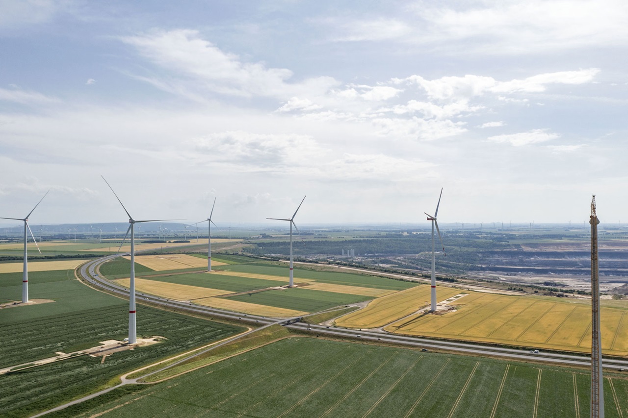 Luftaufnahme von Windkraftanlagen in landwirtschaftlichen Feldern mit bewölktem Himmel und einer kurvigen Straße im Vordergrund.