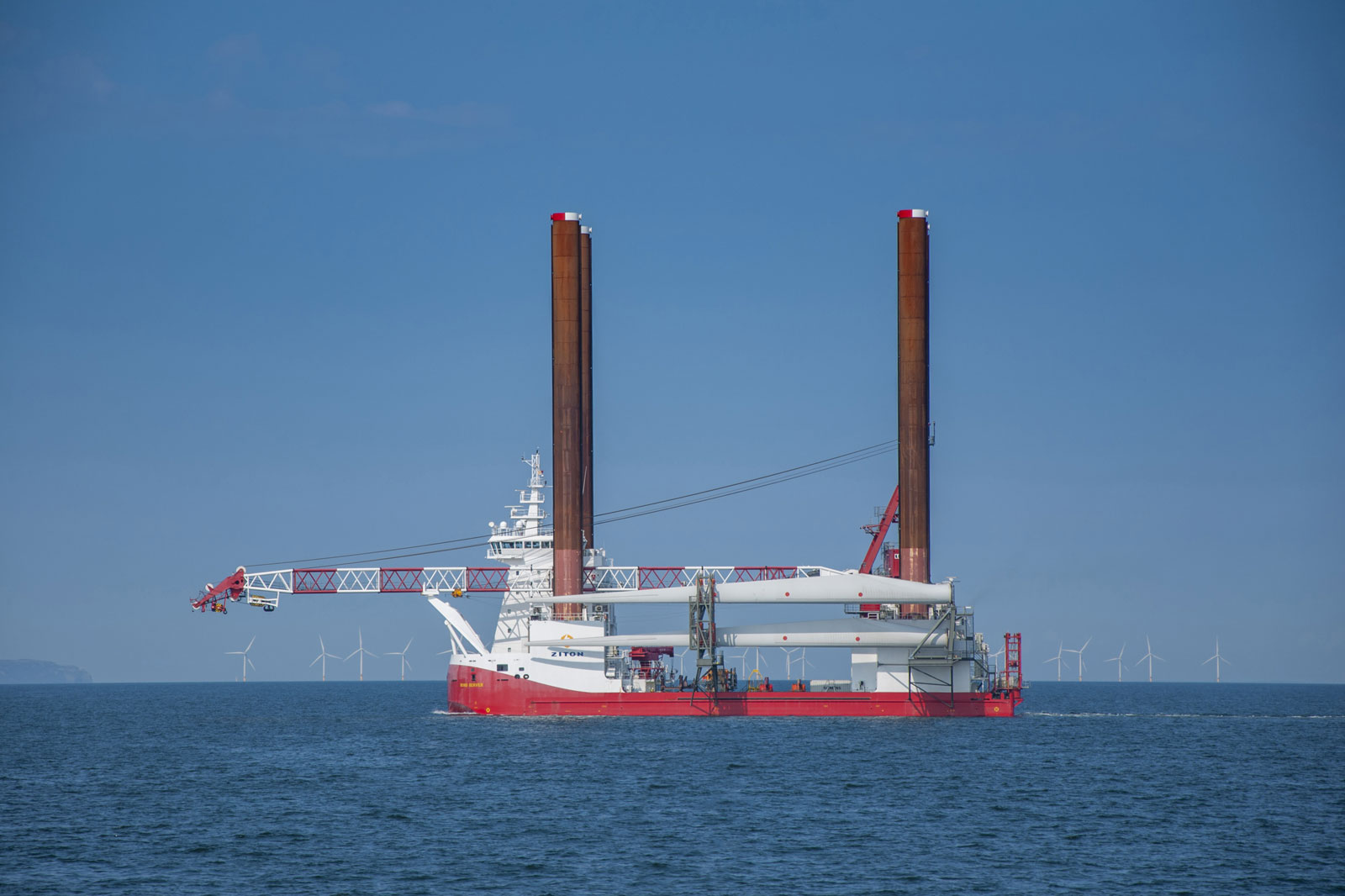 A large offshore construction vessel with two tall brown legs, equipped with a crane, in a clear blue sea, wind turbines in the distance.