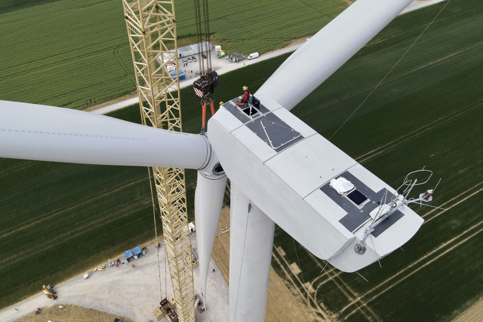 Aerial view of a wind turbine under construction with a worker seated on the platform and a crane in the background.