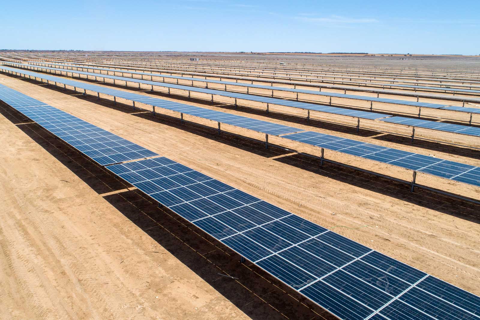 A solar farm with numerous rows of solar panels set on sandy terrain under a clear blue sky.