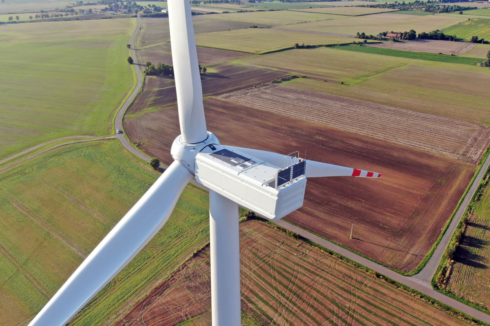Aerial view of a large wind turbine with a white tower and three blades, set against a patchwork of green and brown fields.