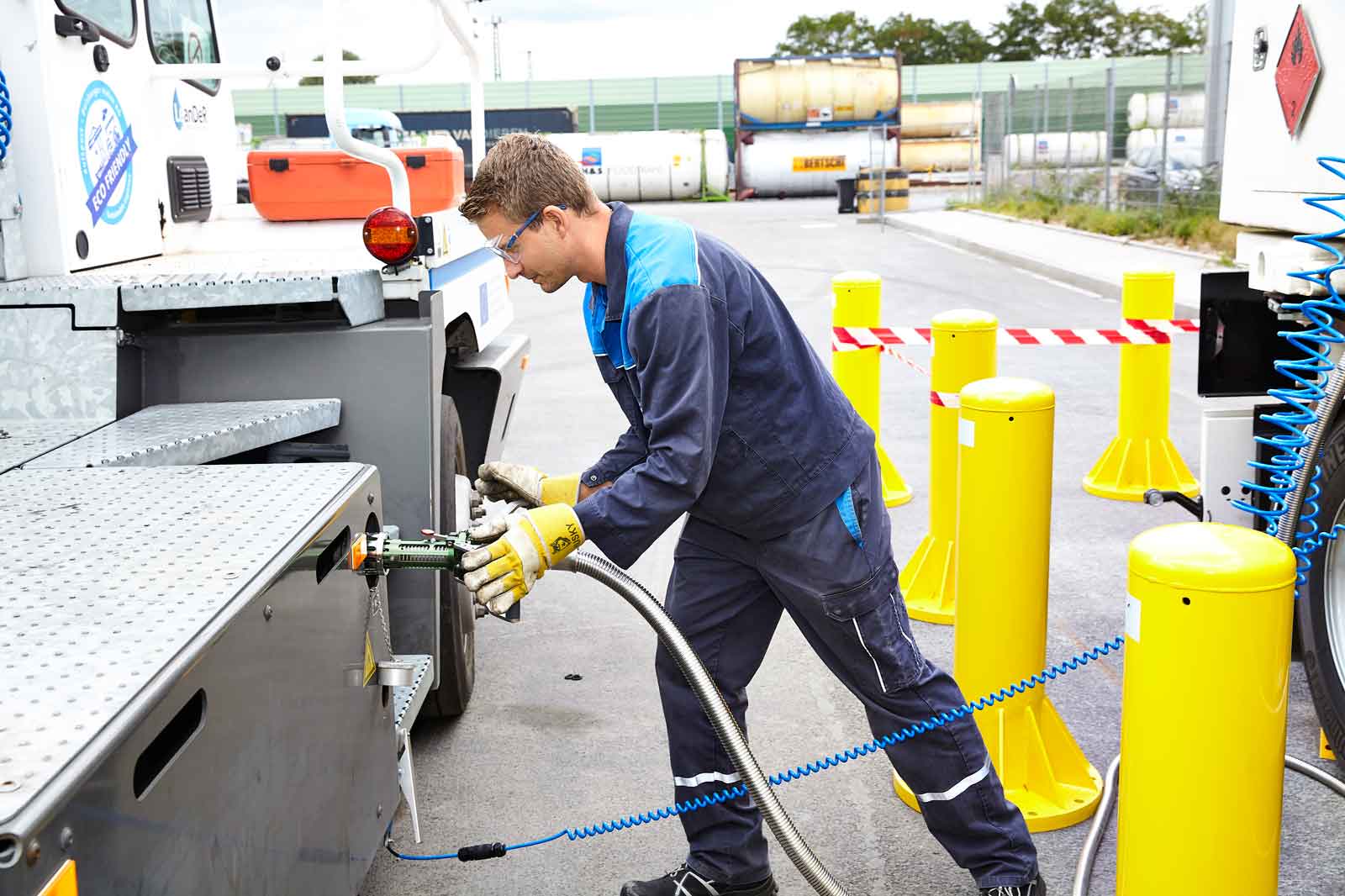Ein Arbeiter in blauen Overall verbindet einen Schlauch mit einem Lastwagen an einer Tankstelle, mit gelben Barrieren im Hintergrund.