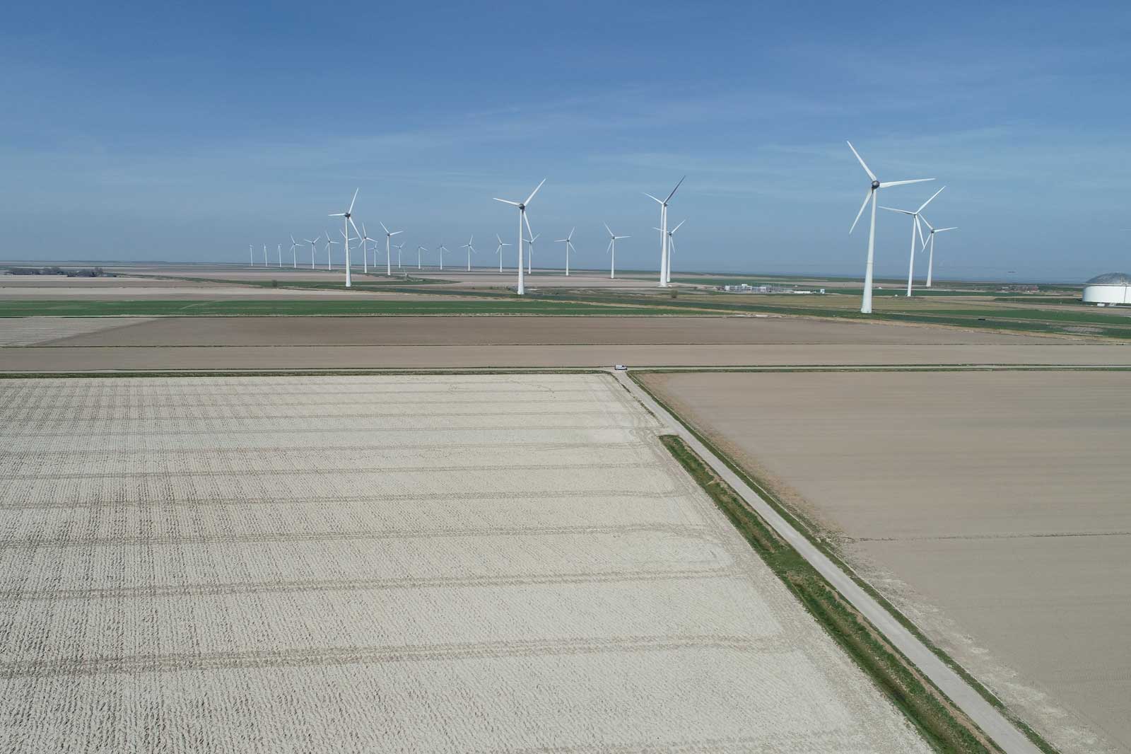 A vast landscape featuring wind turbines. Fields of varying shades of brown and green are visible.