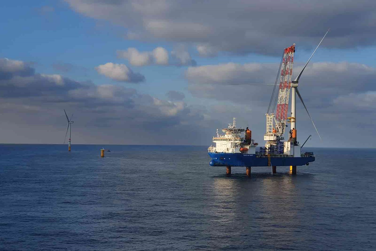 Ein blaues Schiff mit einem Kran ist im Ozean positioniert, im Hintergrund sind Windturbinen unter einem teilweise bewölkten Himmel.