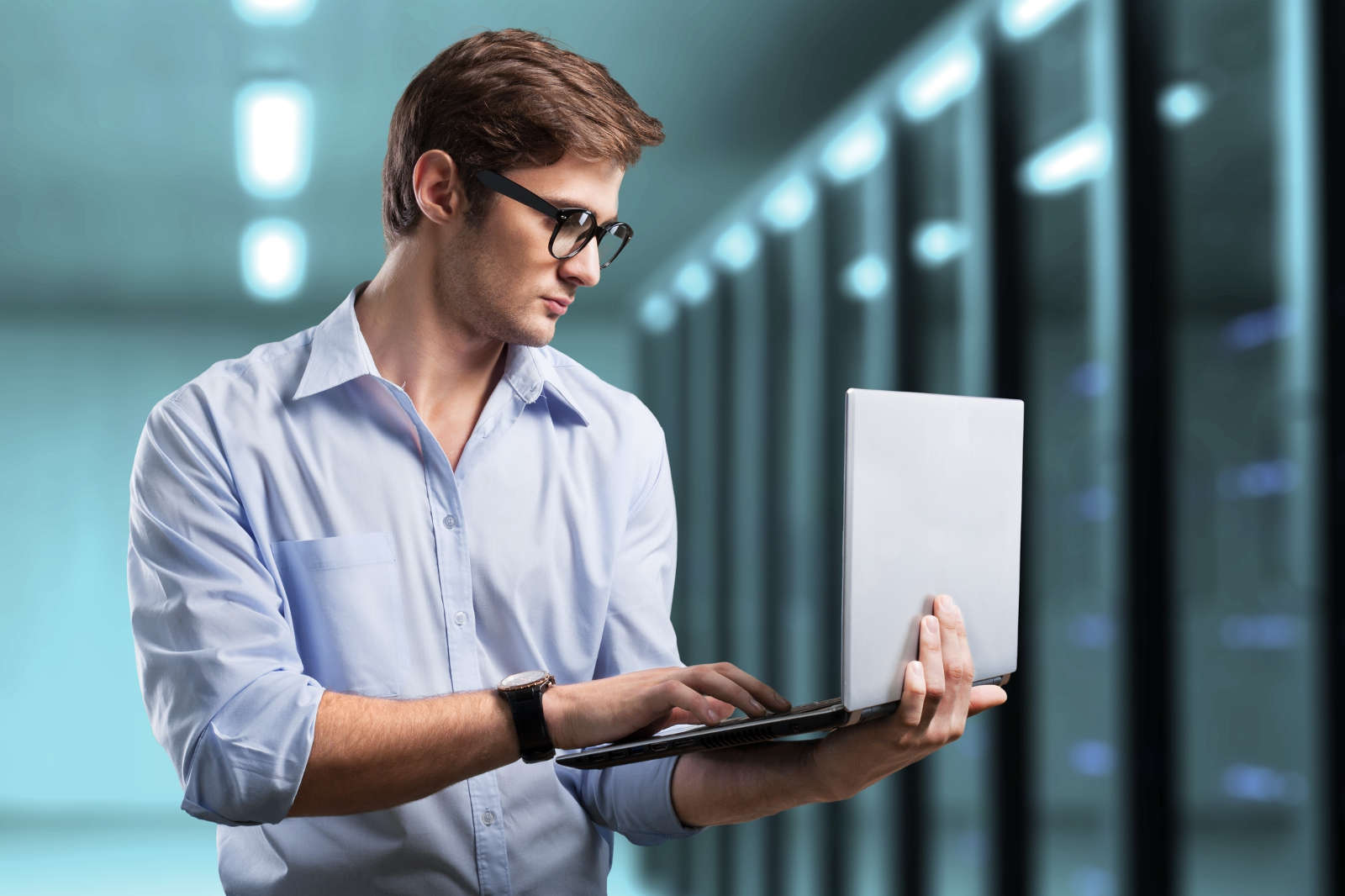 A man in a light blue shirt holds a laptop and a tablet in a modern, well-lit office.