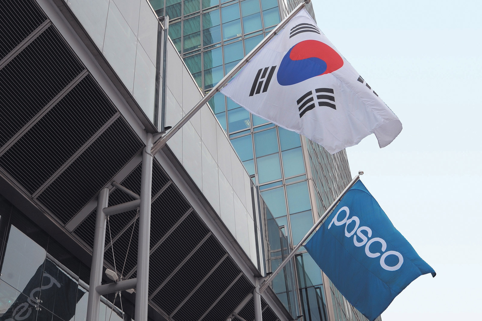 The flags of South Korea and POSCO flutter in front of a modern glass-facade building.