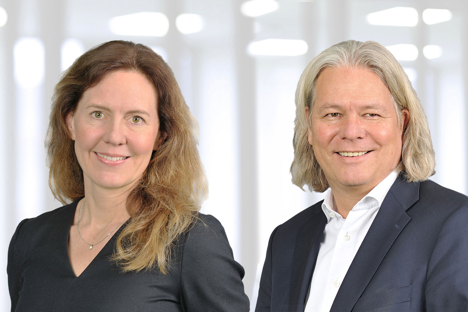 A man and a woman stand side by side. The woman wears a black top, while the man is in a dark suit with a light blue tie.