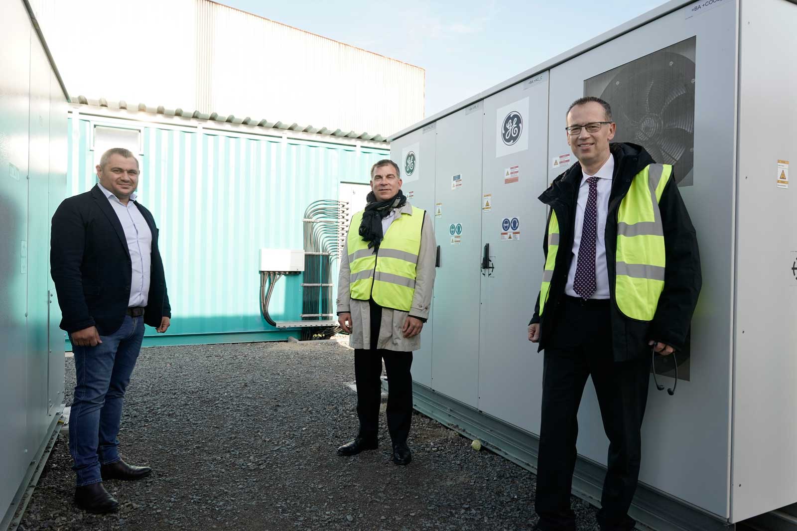 Three men in high-visibility vests stand in front of a building with cooling equipment. The ground is covered with gravel.