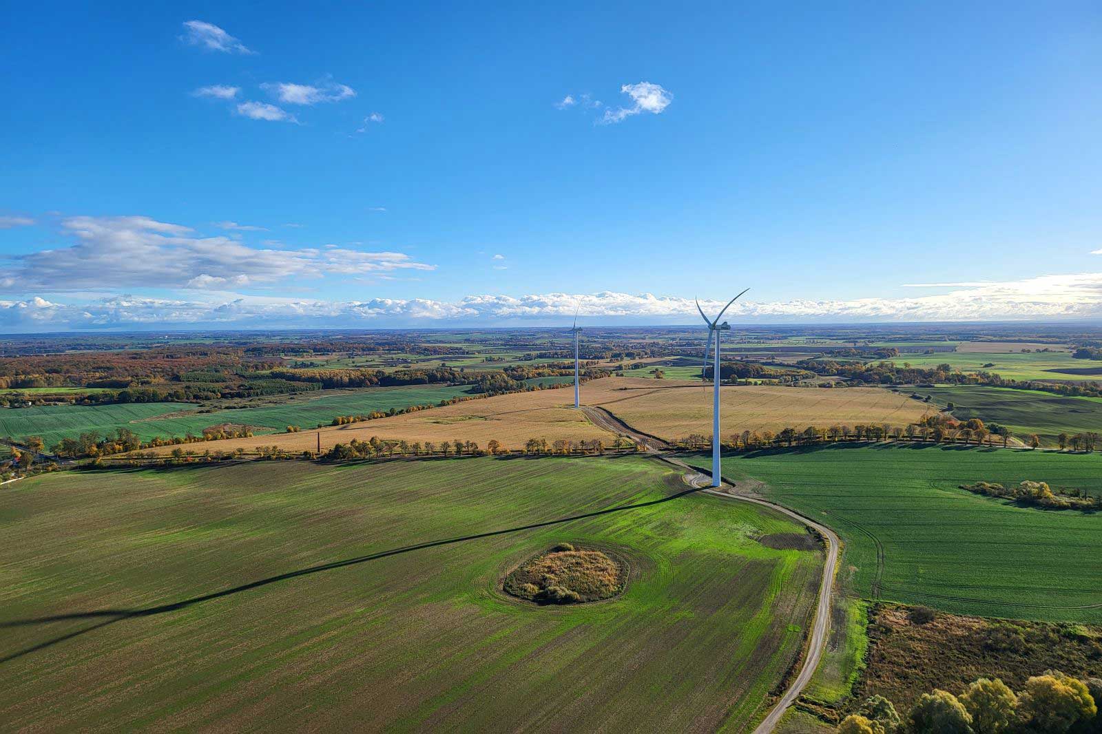 Eine Landschaft mit Windturbinen, grünen Feldern und einem klaren blauen Himmel. Die Bäume zeigen herbstliche Farben.