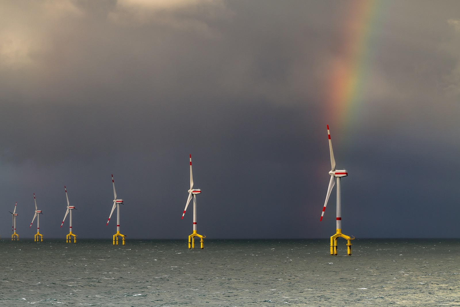Six wind turbines in the sea under a cloudy sky, featuring a rainbow in the background.