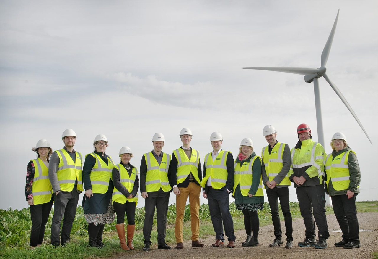 A group of people in safety vests and helmets stand near a wind turbine against a cloudy sky.