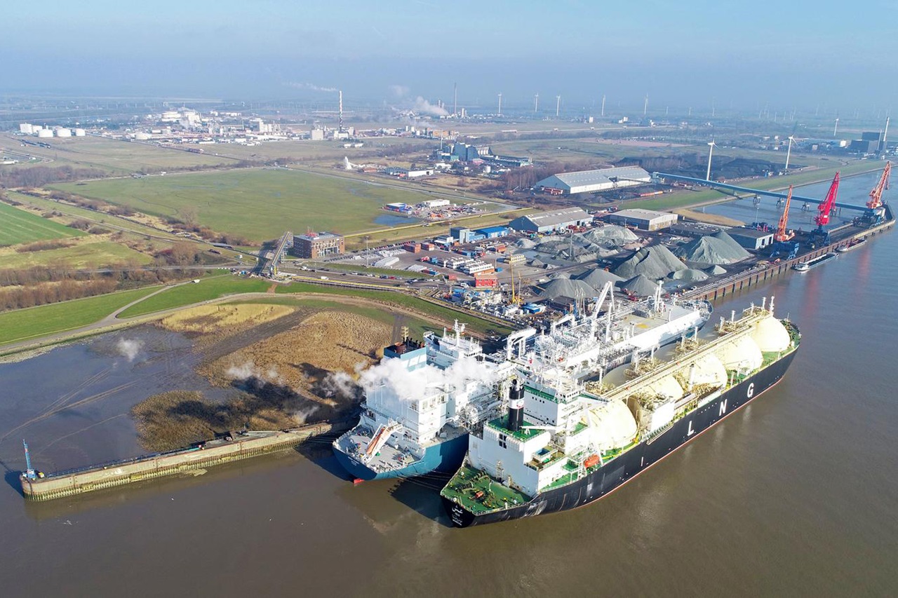 Luftaufnahme eines industriellen Hafenbereichs mit angelegten LNG-Tankern, Lagerhäusern und grünen Feldern im Hintergrund.