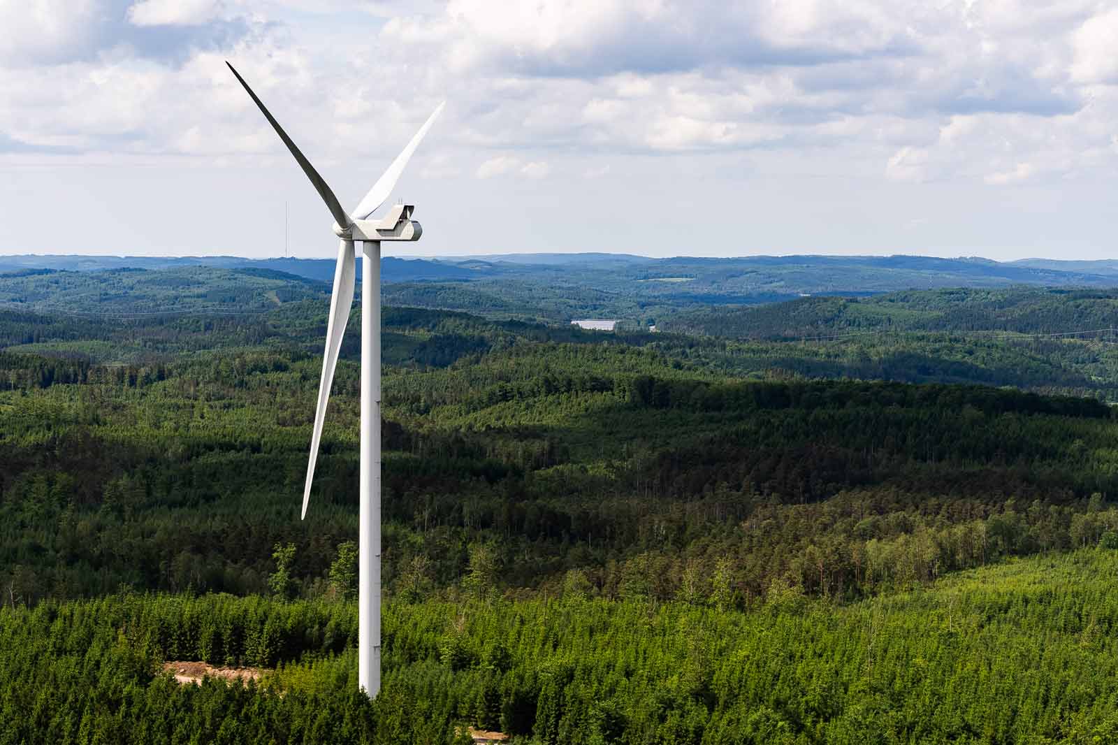 A white wind turbine stands tall amidst a lush green forest, with rolling hills in the background under a partly cloudy sky.