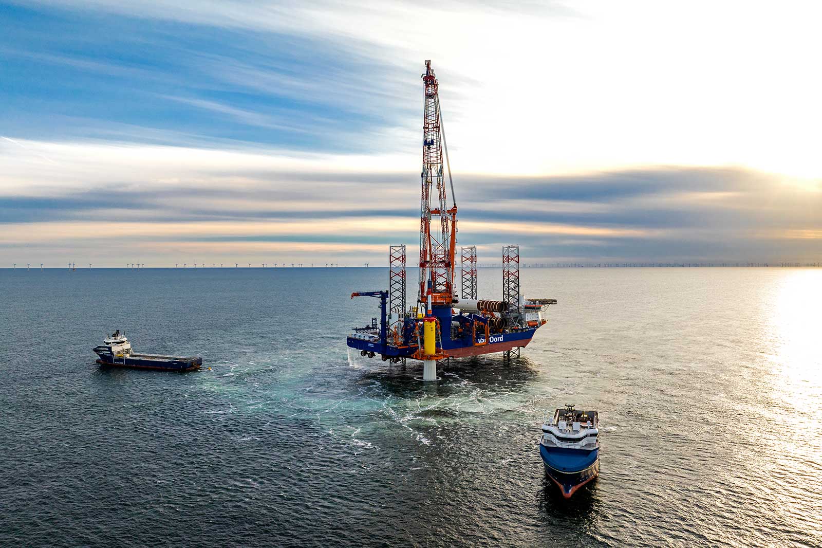 A drilling rig is situated in the sea, surrounded by two ships. The sky is cloudy, and the water reflects the light.