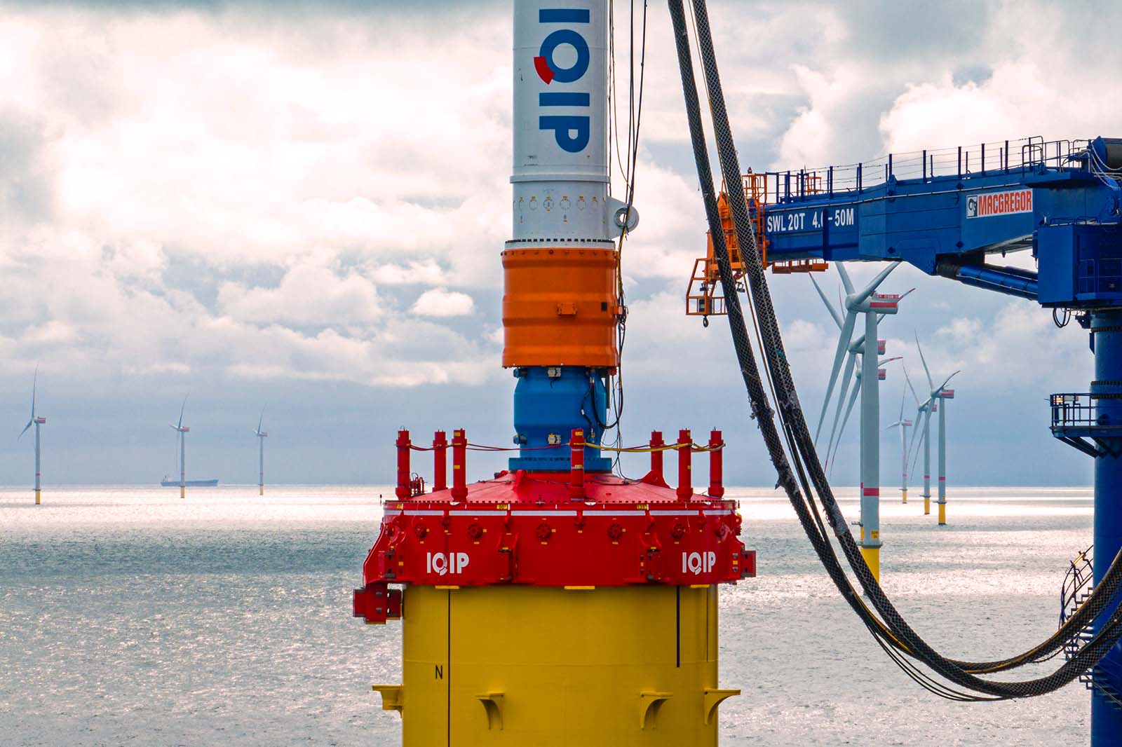 A crane installs a red and yellow offshore wind turbine foundation with thick power cables, with wind turbines on the horizon under a cloudy sky.