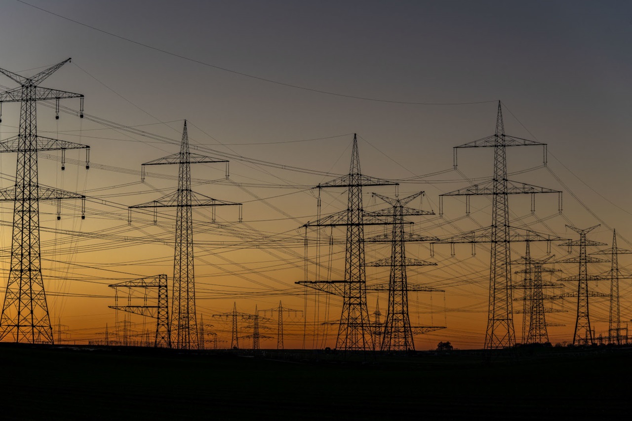 Hochspannungsmasten aus Stahlgitter bilden dunkle Silhouetten in einer flachen Landschaft, zahlreiche Leitungen zeichnen sich vor einem orangenen bis violetten Abendhimmel ab.