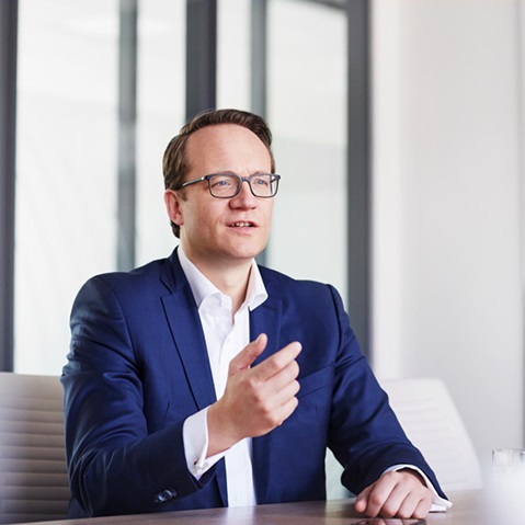 Ein Mann im blauen Anzug sitzt an einem Schreibtisch mit einem Smartphone, Laptop und Computer vor einem lebhaften abstrakten Gemälde. Portrait von Markus Krebber.