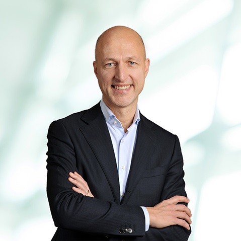 A man in a formal suit stands with crossed arms in front of a blurred, bright background. Portrait of Sven Utermöhlen.