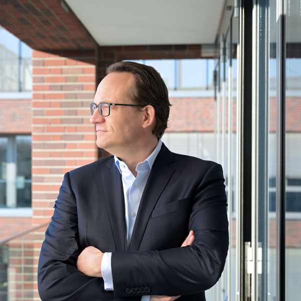 Ein Mann steht mit verschränkten Armen vor großen Fenstern in einem modernen Gebäude. Er trägt einen Anzug. Portrait von Markus Krebber.