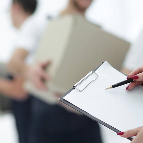 Eine Person hält ein Notizblatt mit einem Stift, während im Hintergrund ein anderer Mensch eine Kiste trägt.