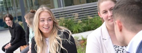 A group of people outdoors engaged in conversation, with plants and modern buildings in the background.
