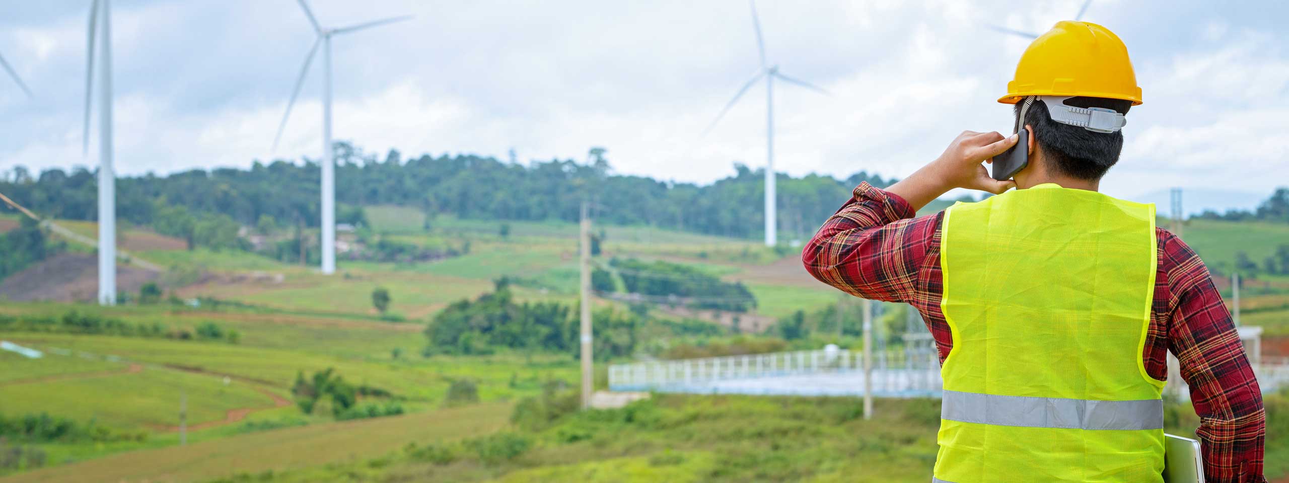 A worker in a yellow hard hat and safety vest is on a mobile phone, overlooking a wind farm in a green landscape.