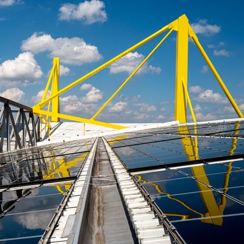 The image features a modern roof structure with yellow supports and reflective solar panels under a blue sky.