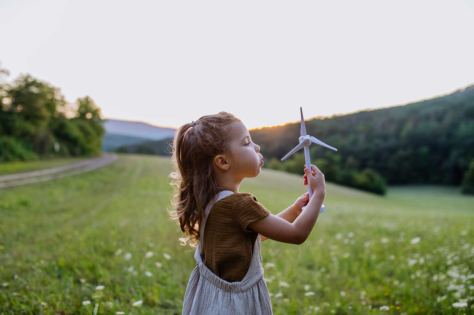 Ein kleines Mädchen hält ein Windrad in der Hand, während es im Freien auf einer Wiese steht, umgeben von Bäumen und sanften Hügeln.