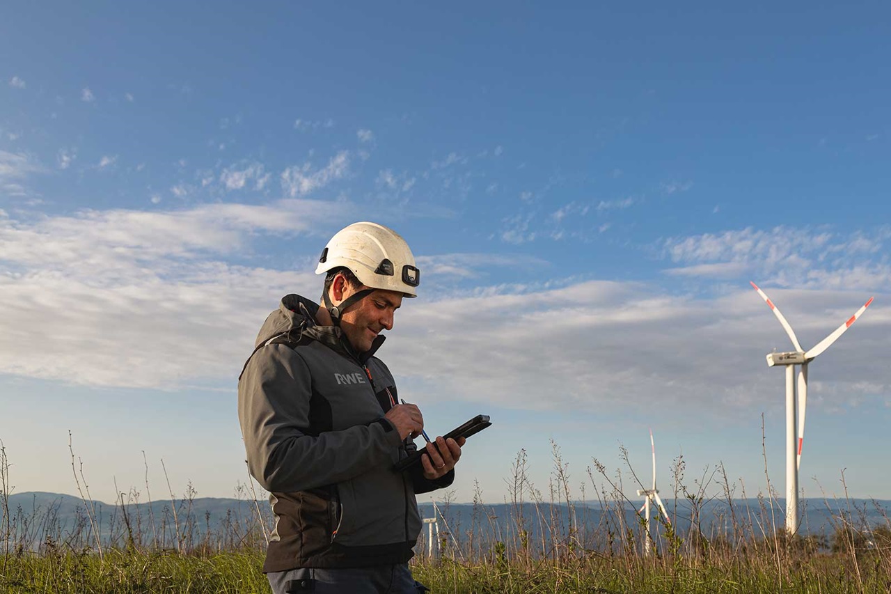 Ein Arbeiter in einem Sicherheitshelm nutzt ein Mobilgerät in einer Windkraftanlage unter einem klaren Himmel.
