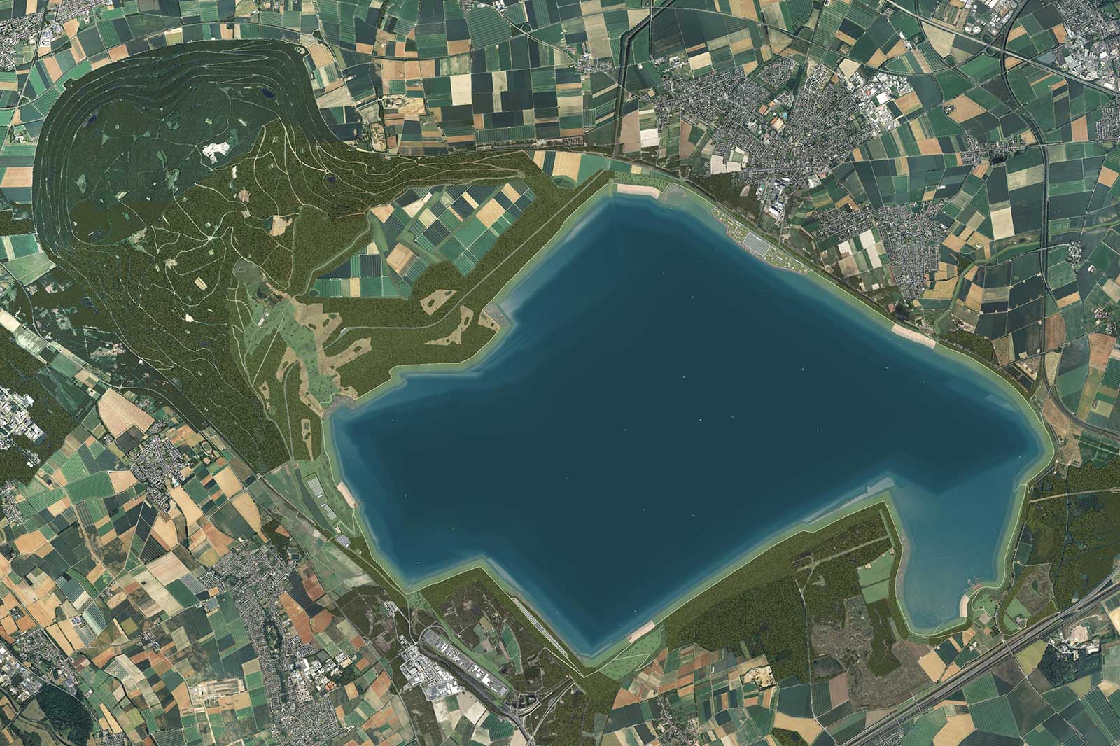 Luftaufnahme eines großen blauen Sees, umgeben von grünen Wäldern und landwirtschaftlichen Flächen, mit einer nahegelegenen Stadt im Hintergrund.
