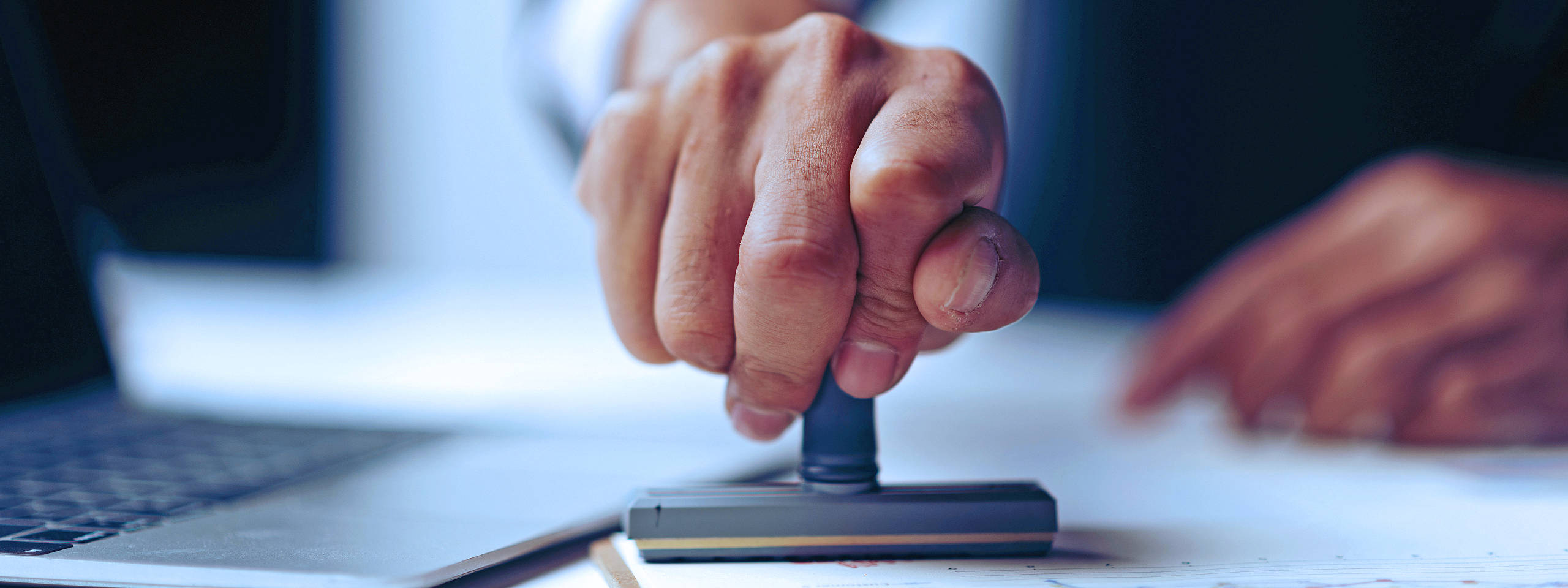 Eine Hand, die einen Gummistempel auf Papier neben einem Laptop drückt, was eine Genehmigung anzeigt.