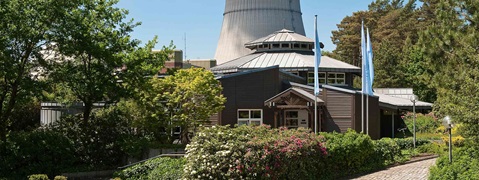 Ein modernes Gebäude mit einem schrägen Dach, umgeben von Grünflächen und blühenden Pflanzen, unter einem klaren blauen Himmel.