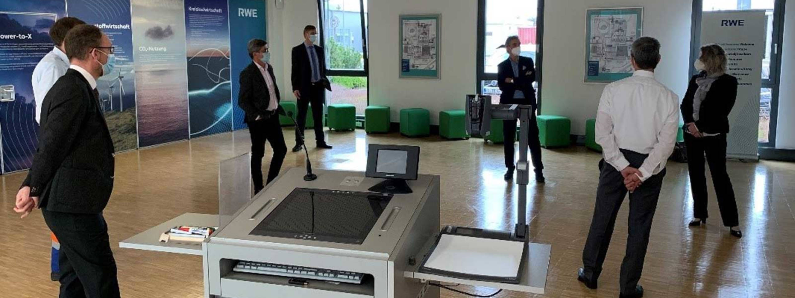 Eine Gruppe von Personen steht in einem modernen Raum mit Präsentationsmaterialien und einem digitalen Arbeitsplatz.