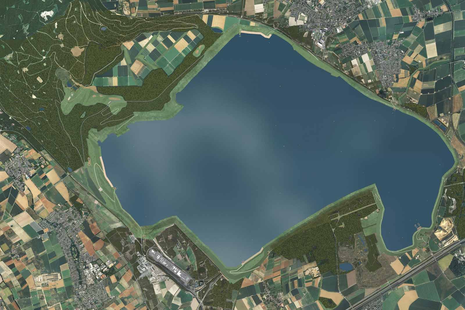 Luftaufnahme eines großen Sees, umgeben von grünen Wäldern und landwirtschaftlichen Feldern. Ein kleiner Flughafen ist in der Nähe des Sees sichtbar.