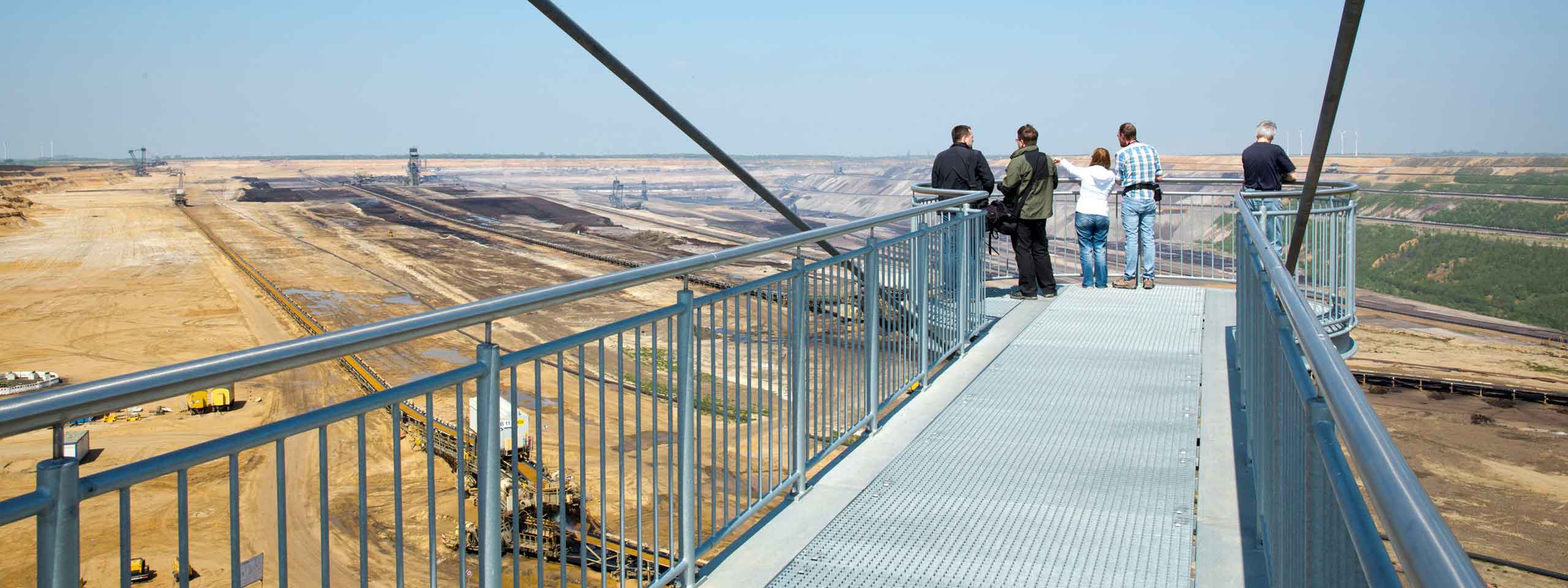Eine Gruppe von Menschen beobachtet eine große Tagebaufläche von einer Aussichtsplattform aus unter klarem Himmel.
