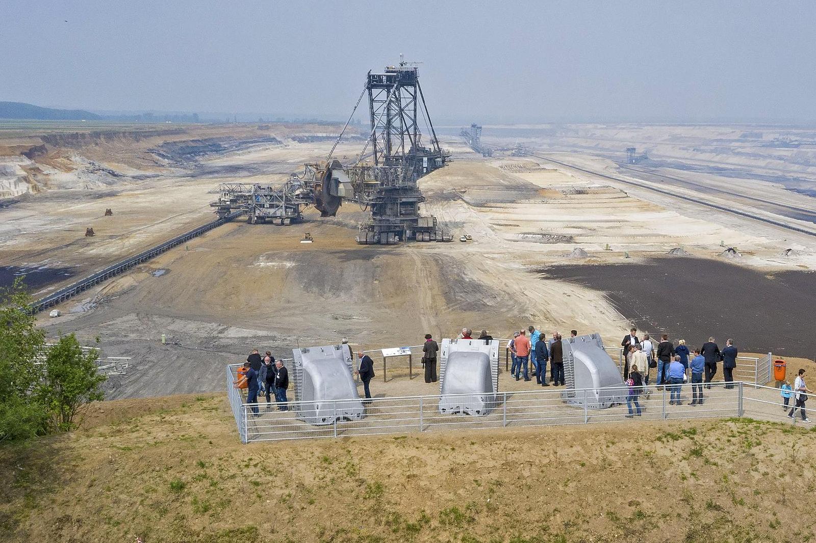 Eine große Abbaumachine steht in einer weitläufigen Landschaft, während Menschen am Rand stehen und beobachten.