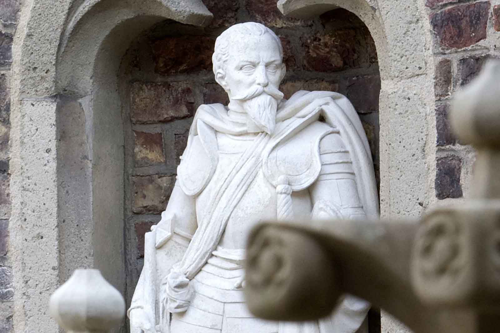 Eine weiße Statue einer Figur in historischer Kleidung steht in einer Nische vor einer Rückenwand aus Ziegeln.