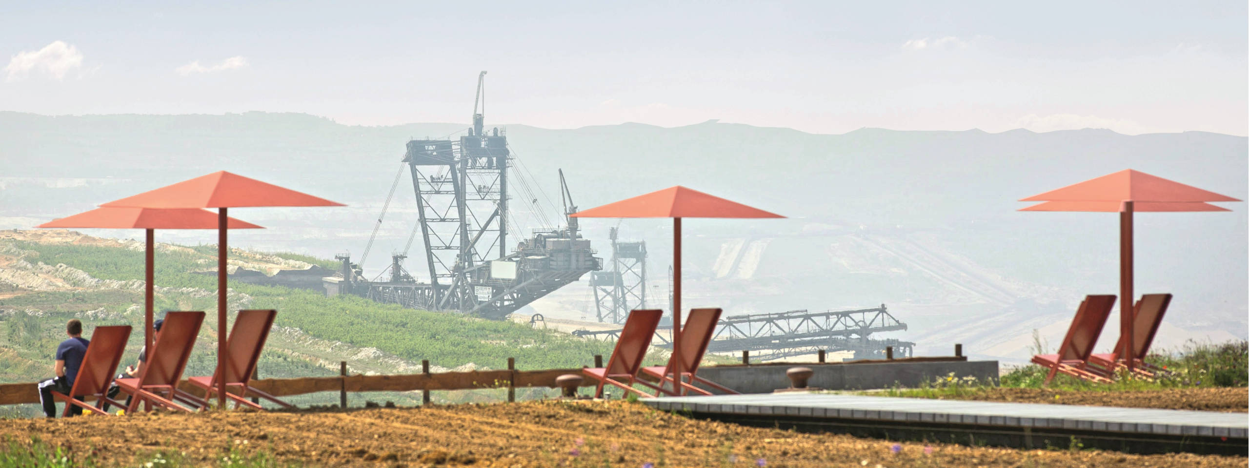 Rote Liegestühle und Sonnenschirme vor einer industriellen Landschaft mit einer großen Maschine im Hintergrund.