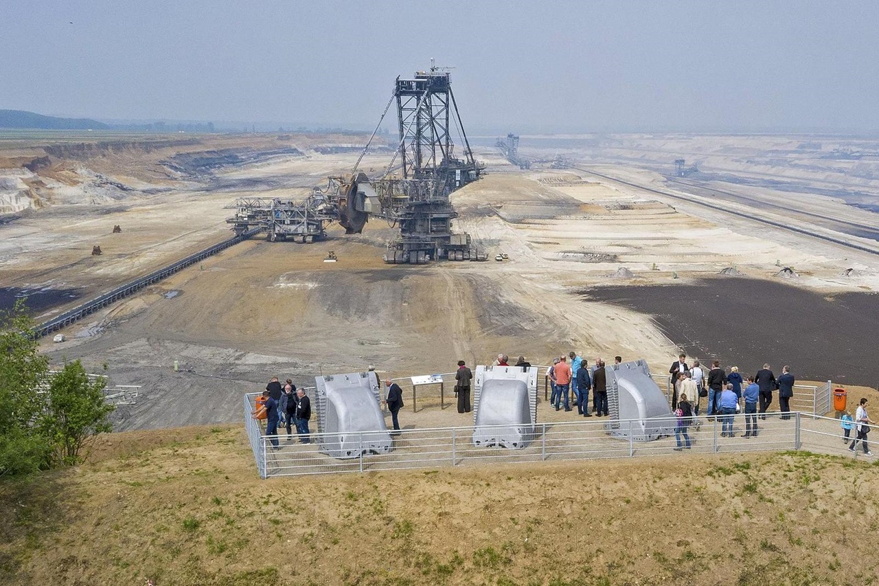 Eine große Abbaumachine steht in einer weitläufigen Landschaft, während Menschen am Rand stehen und beobachten.