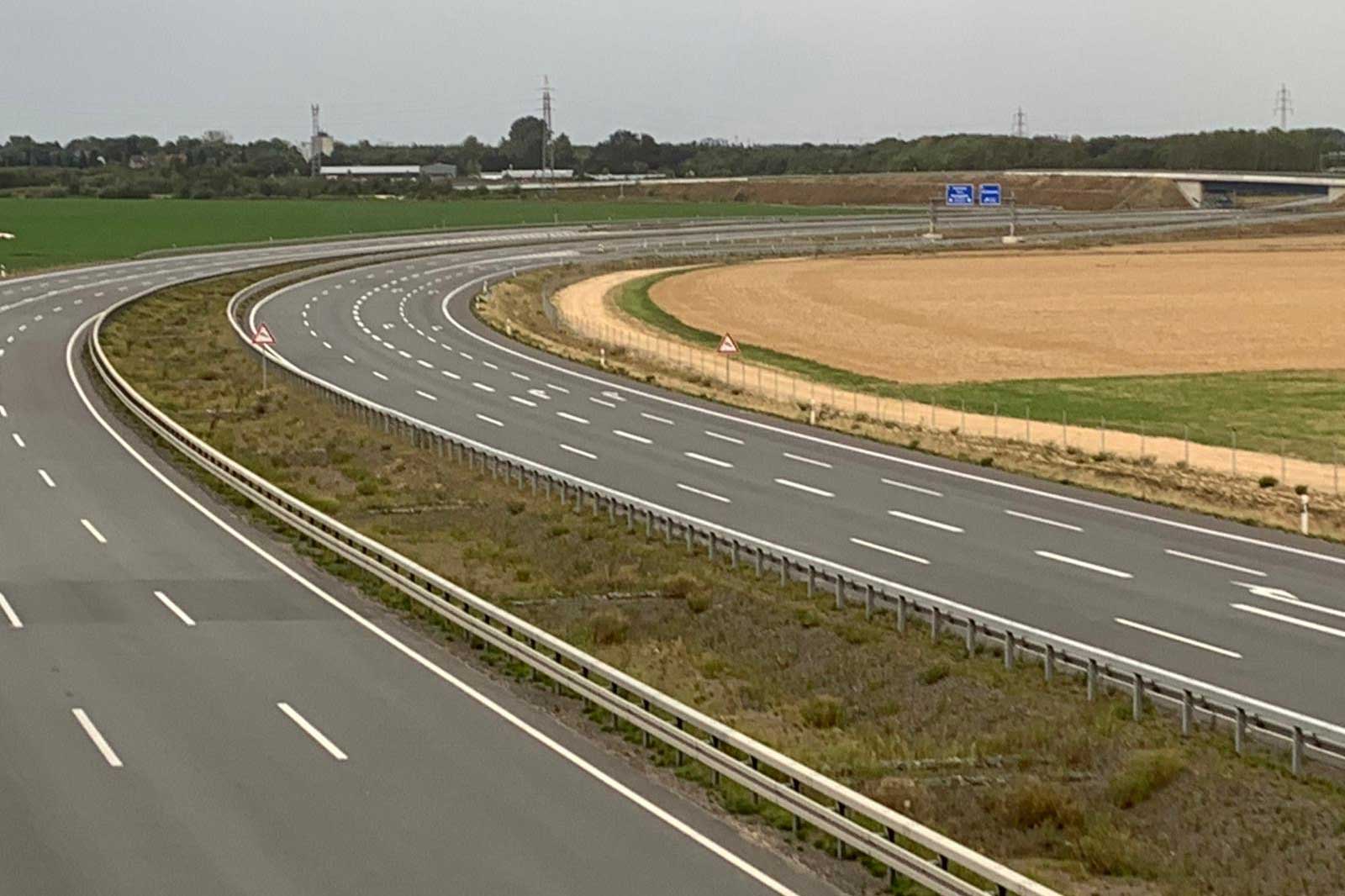 Eine kurvenreiche Autobahn mit mehreren Fahrstreifen, gesäumt von Gras und Feldern unter einem grauen Himmel.