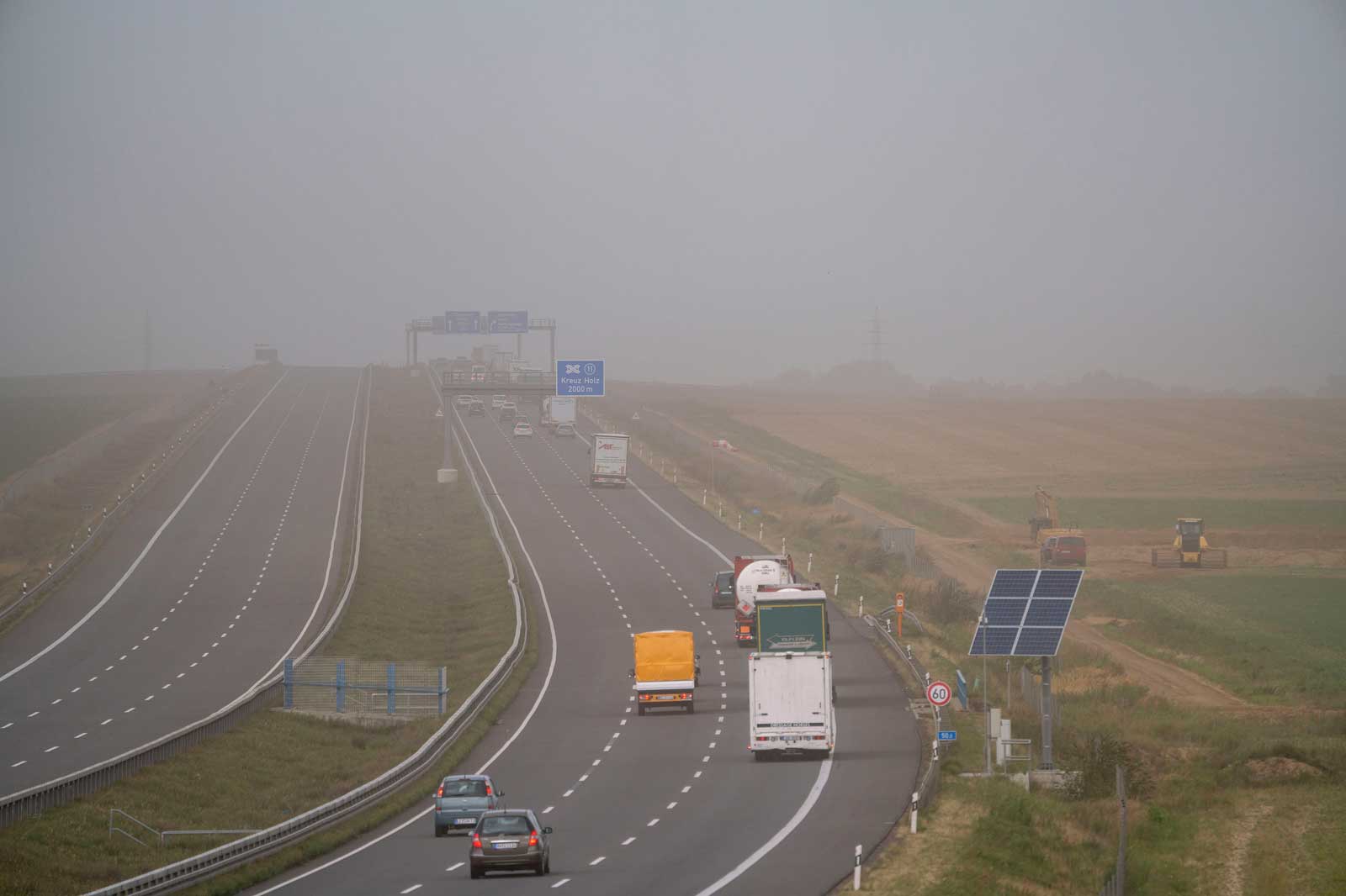 Eine neblige Autobahnansicht mit Autos und Lastwagen, einem Solarpanel am Straßenrand und Baumaschinen auf den Feldern.